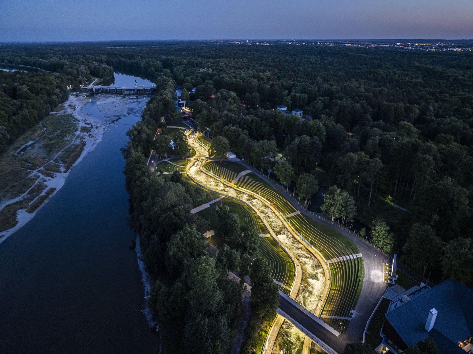 Luftaufnahme des beleuchteten Augsburger Eiskanals in der Abenddämmerung, links der Lech, ringsum Wald