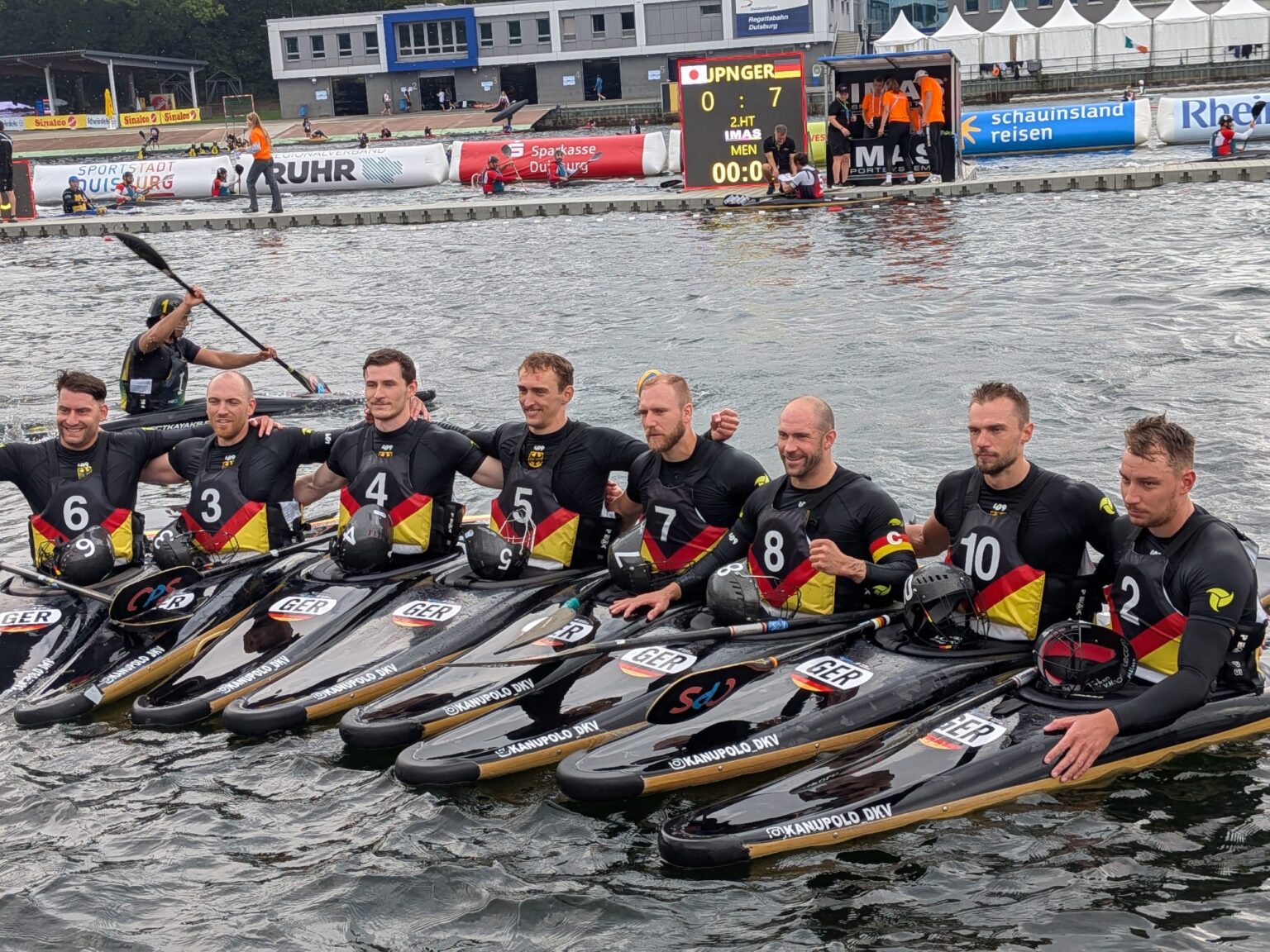 Die deutschen Kanu-Polo-Herren sitzen Arm in Arm in ihren Booten nebeneinander, im Hintergrund zeigt die Anzeigetafel das 7:0 gegen Japan.