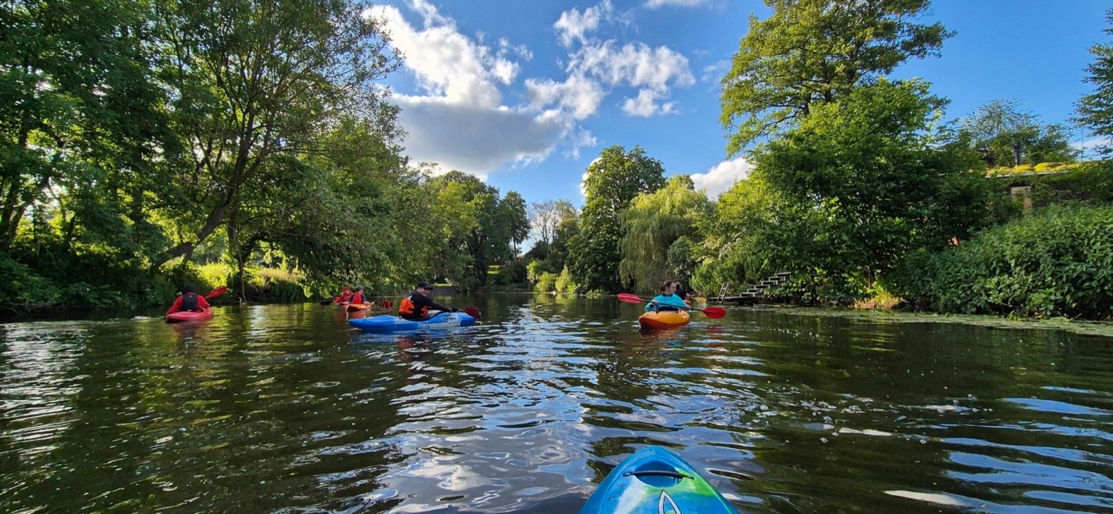 Mehrere Paddler in bunten Kajaks auf der Werse, an beiden Ufern hohe Bäume, darüber blauer Himmel mit Wolken.