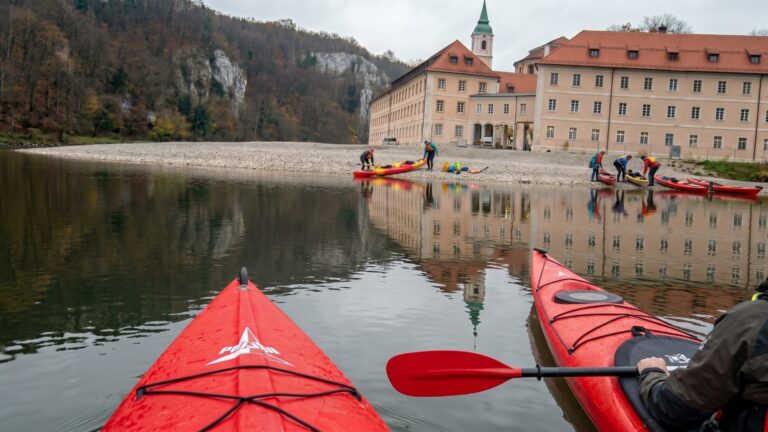 Blick über den Bug eines roten Seekajaks auf eine Kiesbank an der Donau, wo Paddler ihre Boote beladen, dahinter eine Klosteranlage und bewaldete Felshänge.