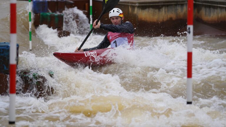 Stefan Hengst mit Startnummer 1 im roten Kajak im Weißwasser zwischen den Torstangen im Kanupark Markkleeberg.