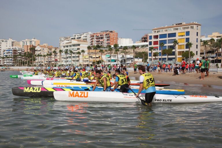 Aufgereihte Surfskis im flachen Wasser vor dem Strand von La Vila Joiosa, die Paddler mit Startnummern warten neben ihren Booten auf das Startsignal.